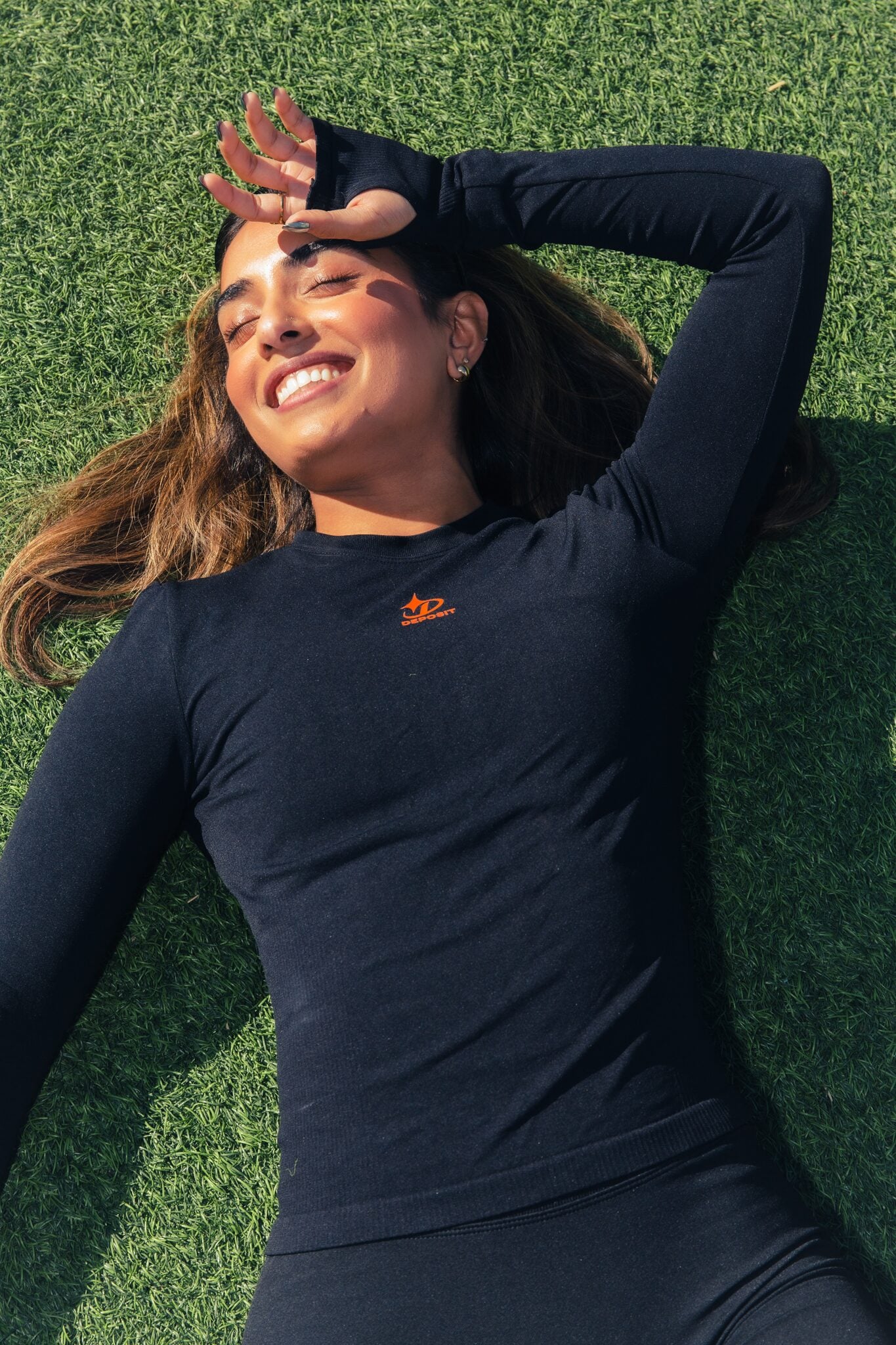 Woman laughing on turf, hands framing her face, wearing a long-sleeve compression top.